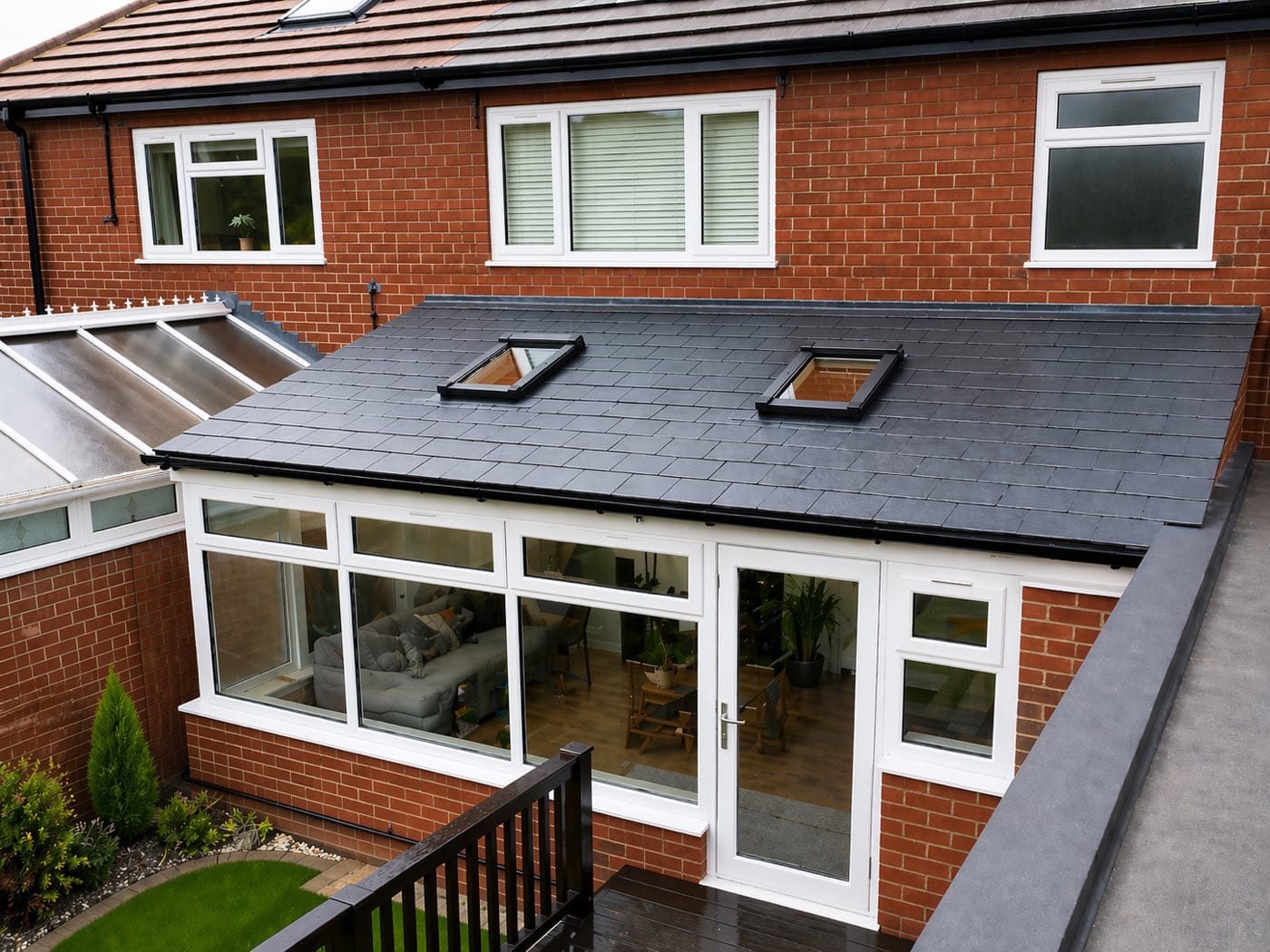 An elevated view of a conservatory with a new slate grey warm roof and two skylights, the old polycarbonate roof still visible alongside it
