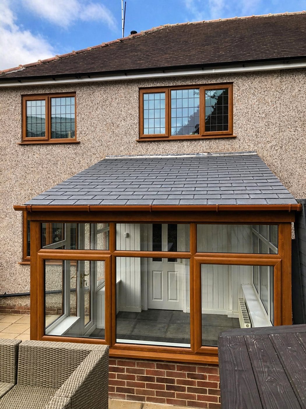 A conservatory with a slate grey tiled warm roof and golden oak frames, fitted to a rendered house