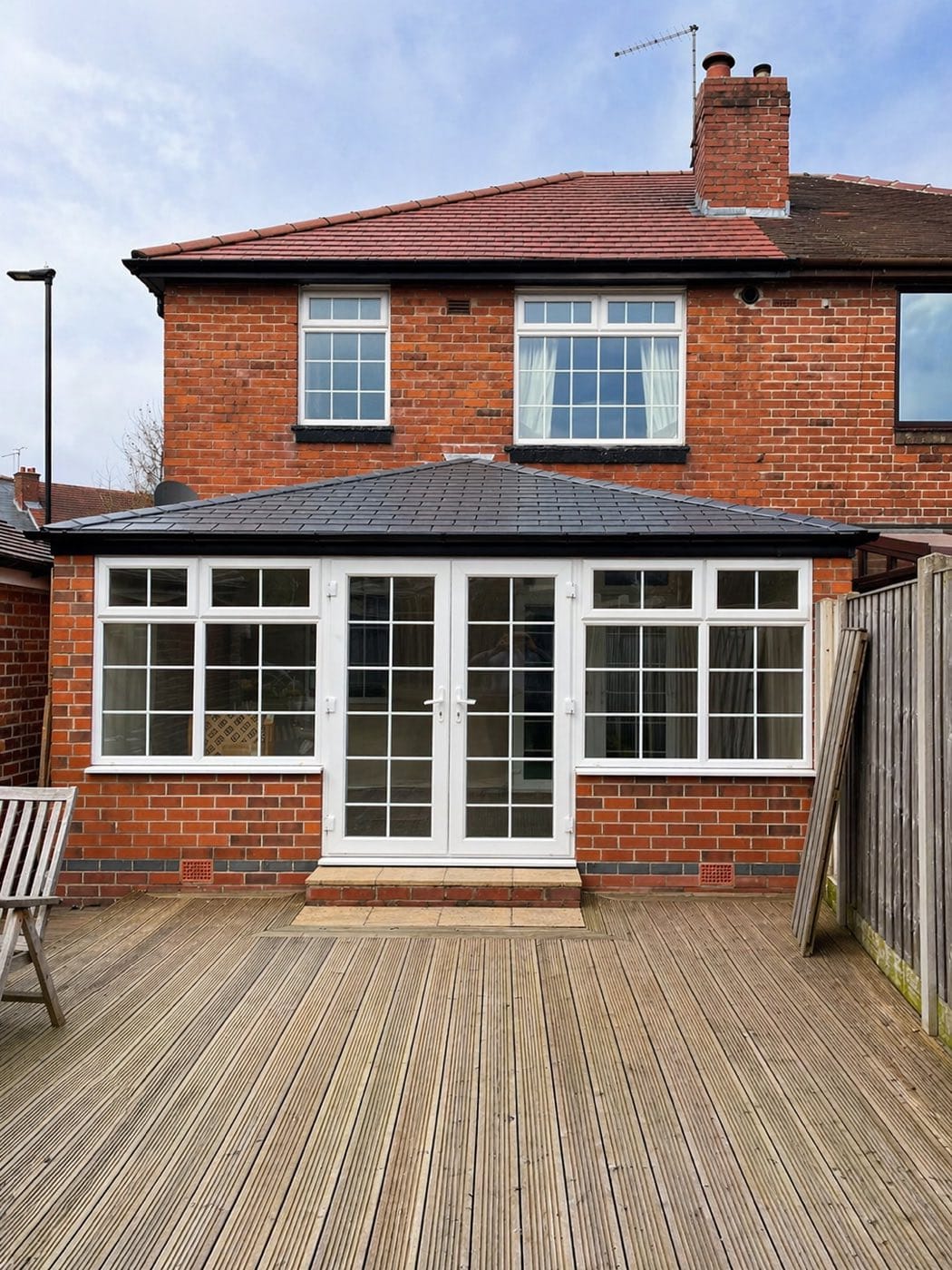 A conservatory with a dark grey tiled warm roof and French doors, on a red brick semi with decking in front