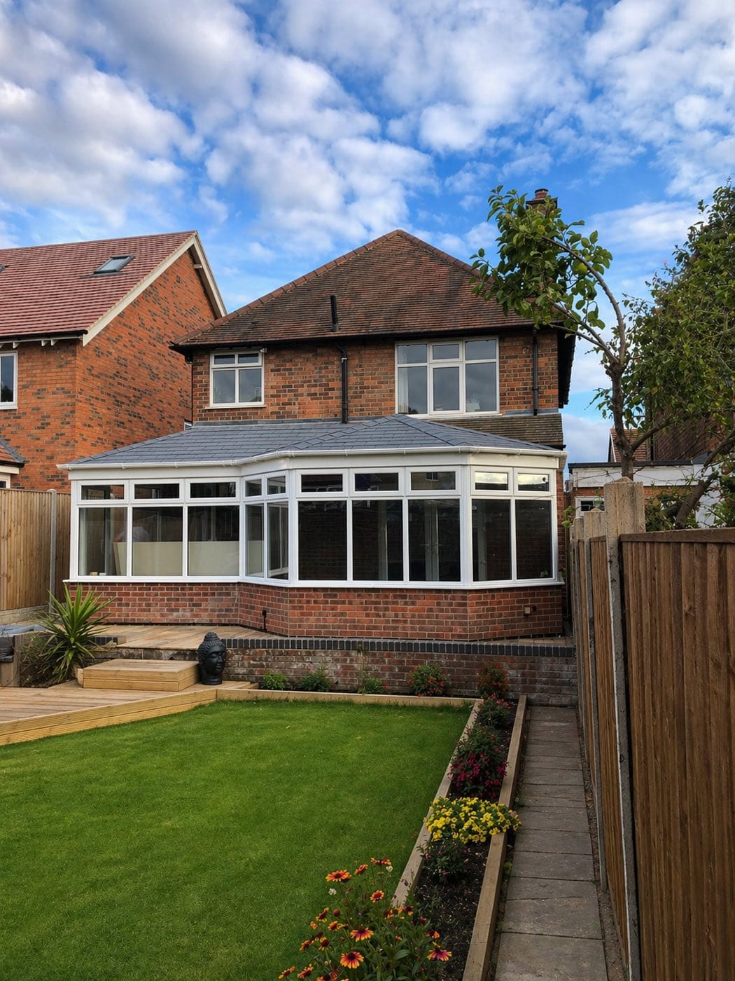 A Victorian style conservatory with a grey tiled warm roof and white frames, on a red brick detached house