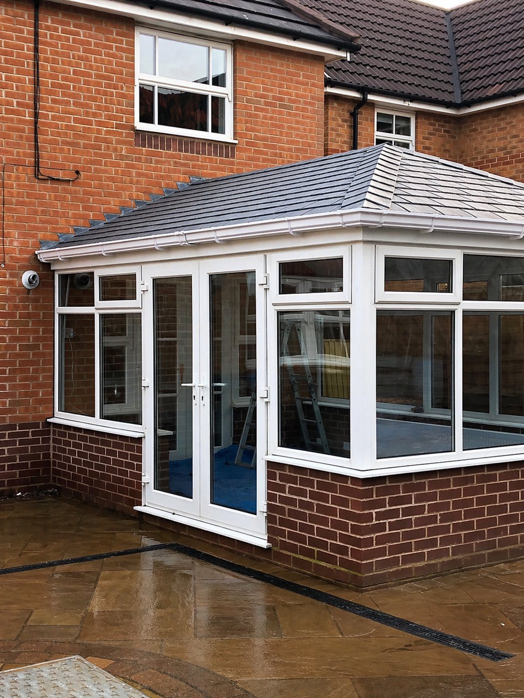 A conservatory with a grey tiled warm roof and French doors, fitted against a red brick house